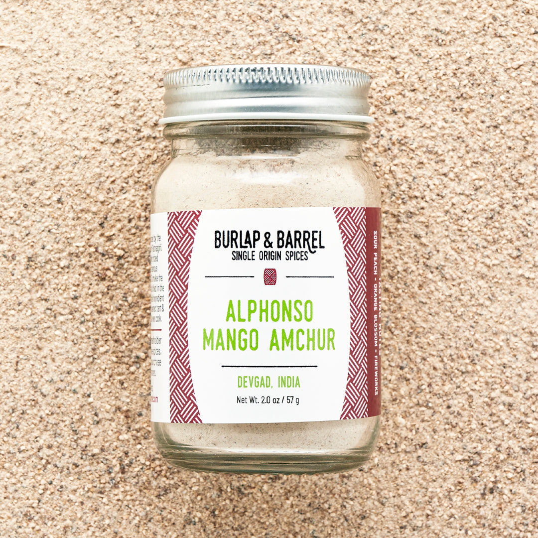 A jar of Alphonso Mango Amchur spice on a background of loose amchur powder with the label 'Burlap & Barrel - Alphonso Mango Amchur