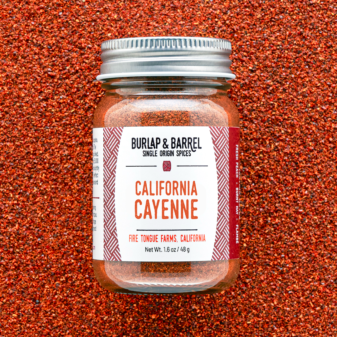 A glass jar of Burlap & Barrel California Cayenne spice blend, filled with orange-red seasoning and topped with a silver metal lid, set against a textured background of loose orange-red cayenne pepper.