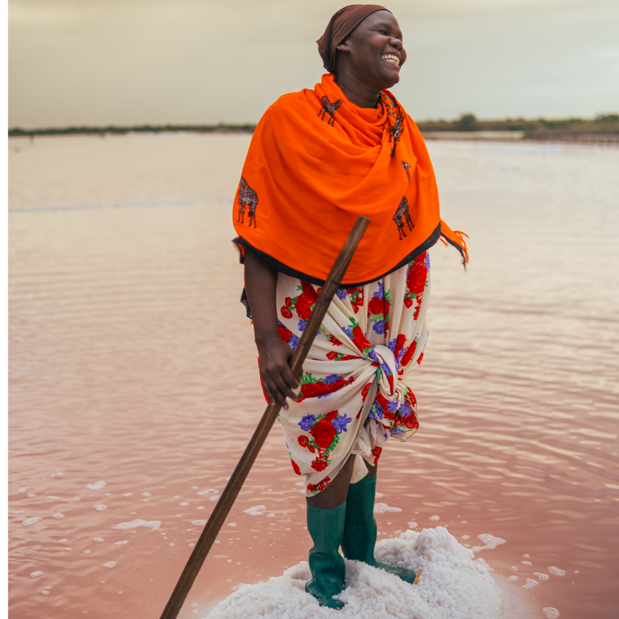 Partner farmer with an orange shawl smiling atop of a pile of salt. Surrounding her is water and she is holding a long stick.