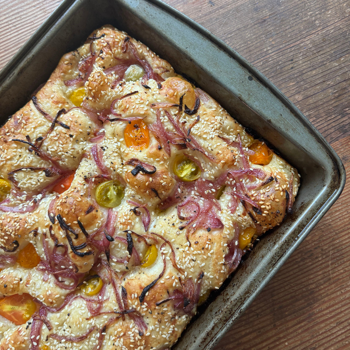 A baked focaccia bread in a dark metal baking pan on a wooden table. The focaccia is topped with sesame seeds, sliced red onions, halved cherry tomatoes, and sliced green olives.