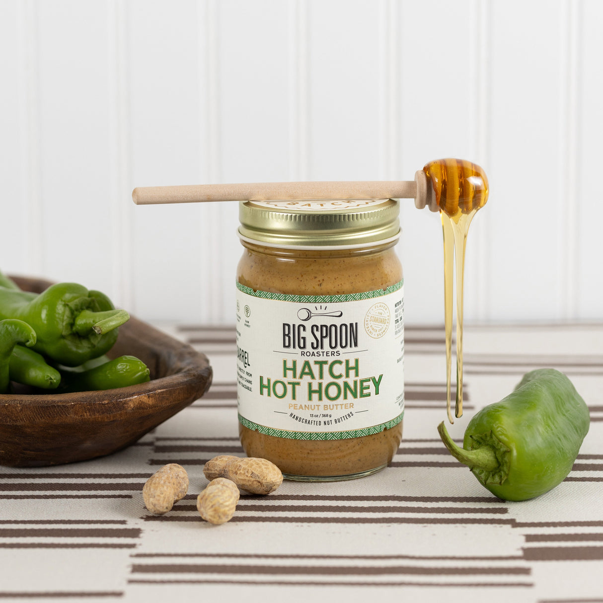 A jar of Big Spoon Farms Hatch Hot Honey sits on a striped tablecloth with a wooden honey dipper dripping honey, green peppers, peanuts, and a bowl of peppers in the background.