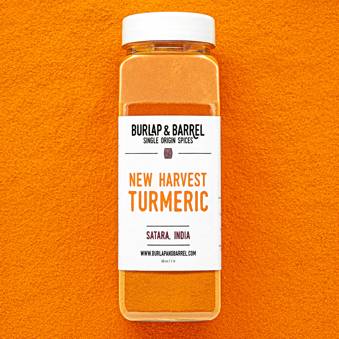 Plastic container of New Harvest Turmeric from Burlap & Barrel with a white lid on a background of loose turmeric powder.