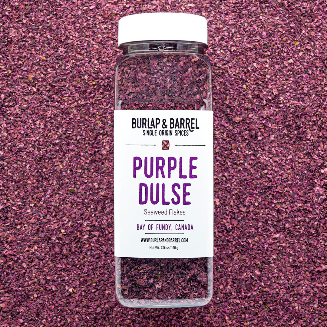 A container of Purple Dulse Flakes by Burlap & Barrel, sustainably hand-harvested from the Bay of Fundy, Canada, displayed on a background of dulse flakes.