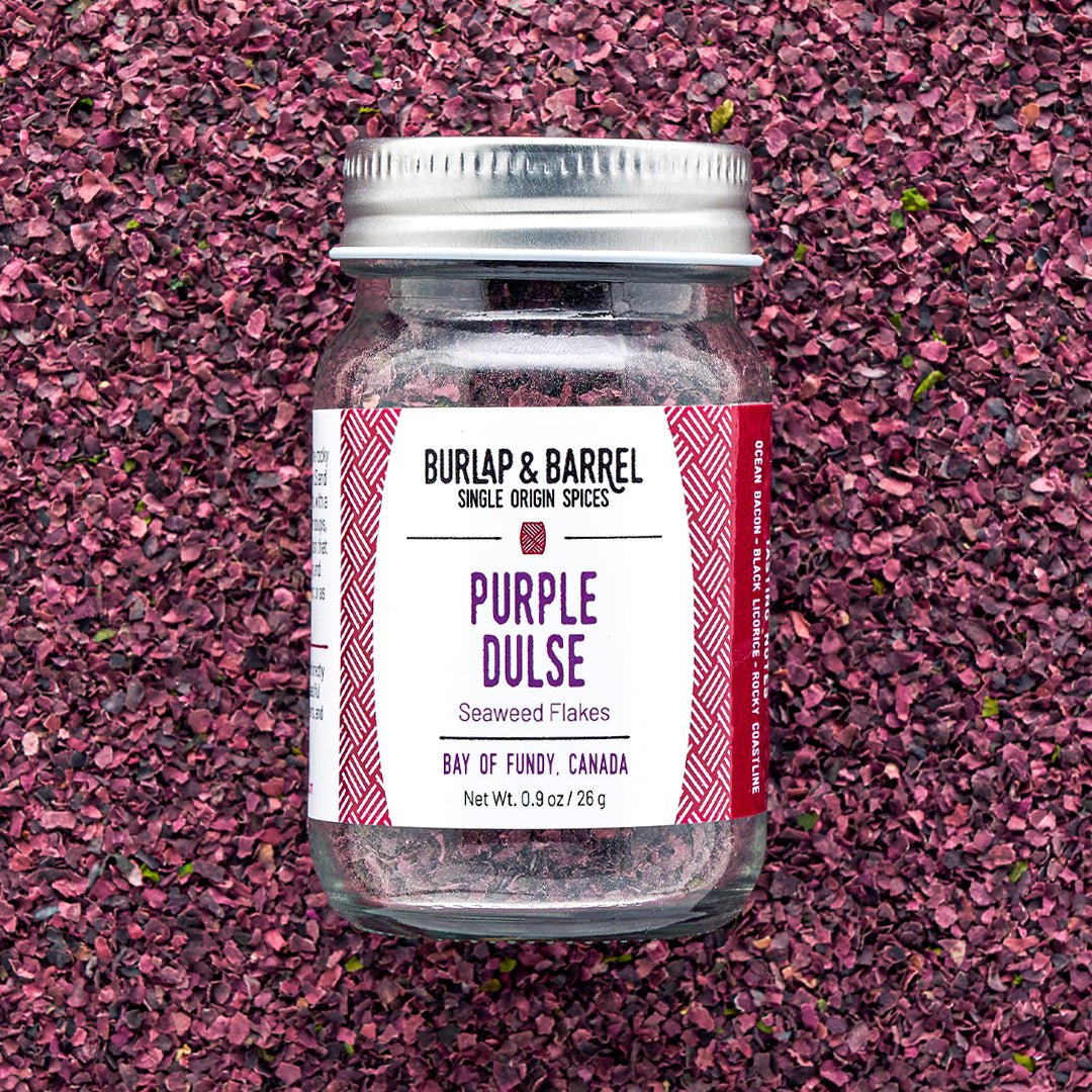 A jar of Purple Dulse Flakes by Burlap & Barrel, sustainably hand-harvested from the Bay of Fundy, Canada, displayed on a background of dulse flakes.