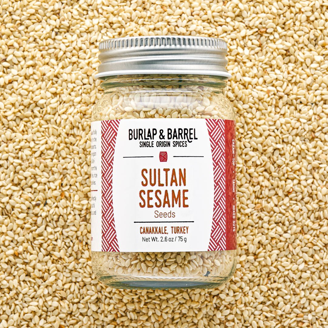 A glass jar of Sultan Sesame seeds by Burlap & Barrel, filled with sesame seeds and topped with a silver metal lid, set against a background of loose sesame seeds. The label features a white background with red patterned borders and brown text.