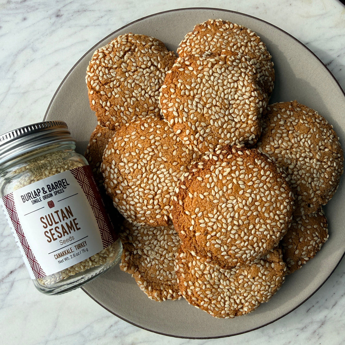 A plate of golden-brown sugar cookies heavily topped with white sesame seeds sits on a white marble surface next to a glass jar of Burlap & Barrel Seasoned Sesame.