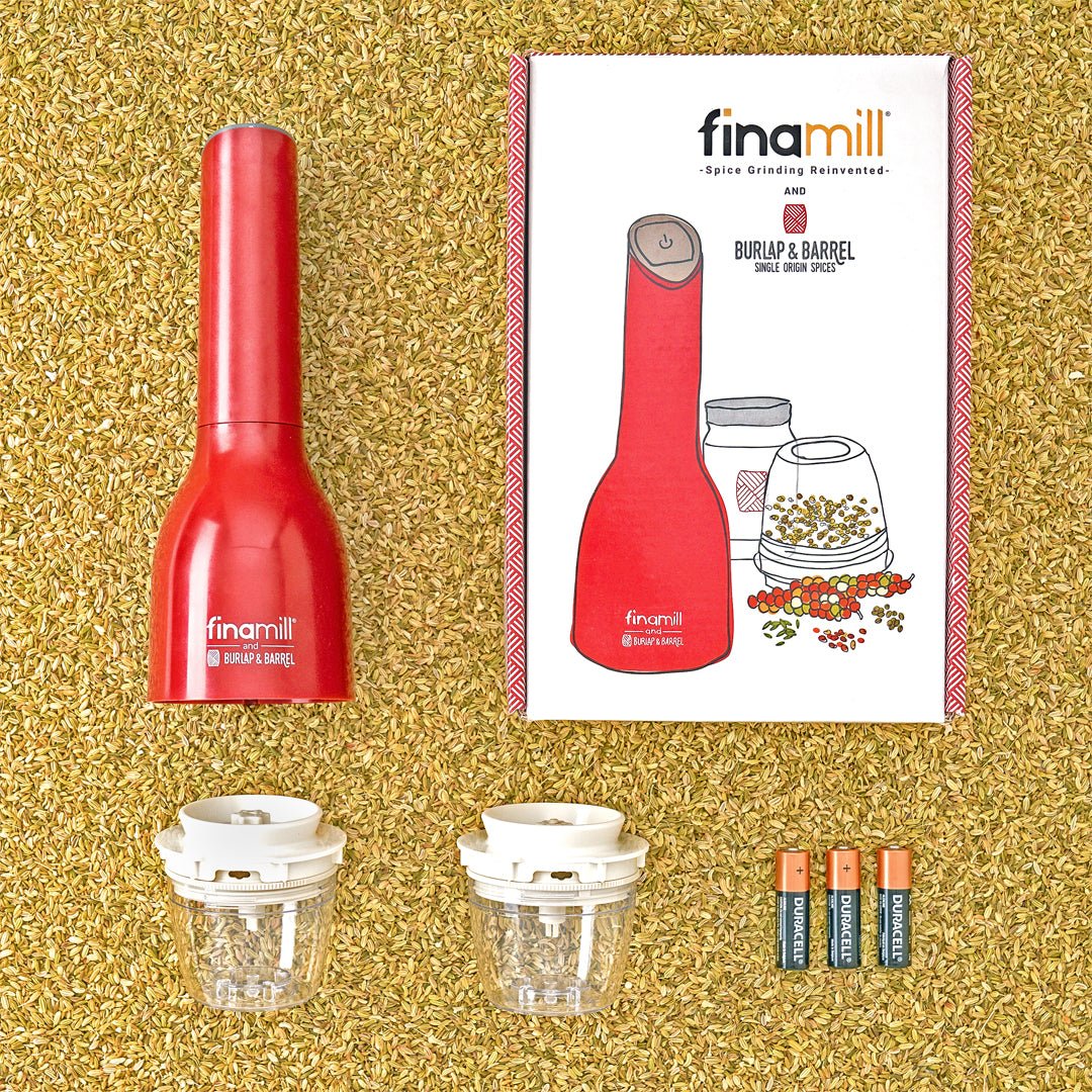 Finamill automatic grinder with the box it comes in, two removable grinding pods and 3 batteries on a bed of loose fennel seeds