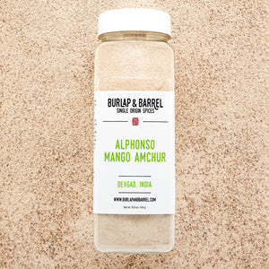 A jar of Alphonso Mango Amchur spice on a background of loose amchur with the label 'BURLAP & BARREL' and 'Alphonso Mango Amchur