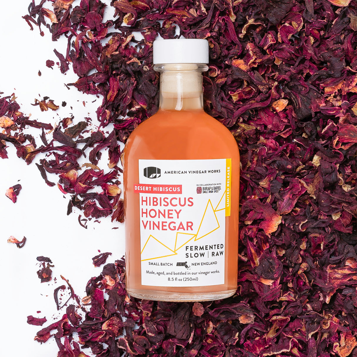 A bottle of Honey Hibiscus Vinegar with a label that reads 'Robert Hibiscus Honey Vinegar, American Craft Vinegar', surrounded by hibiscus flower petals.
