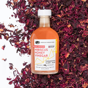 A bottle of Honey Hibiscus Vinegar with a label that reads 'Robert Hibiscus Honey Vinegar, American Craft Vinegar', surrounded by hibiscus flower petals.