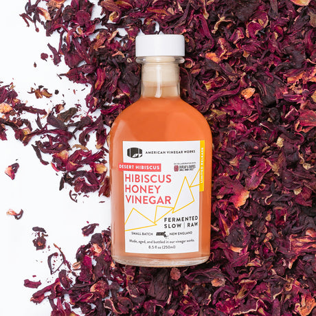 A bottle of Honey Hibiscus Vinegar with a label that reads 'Robert Hibiscus Honey Vinegar, American Craft Vinegar', surrounded by hibiscus flower petals.