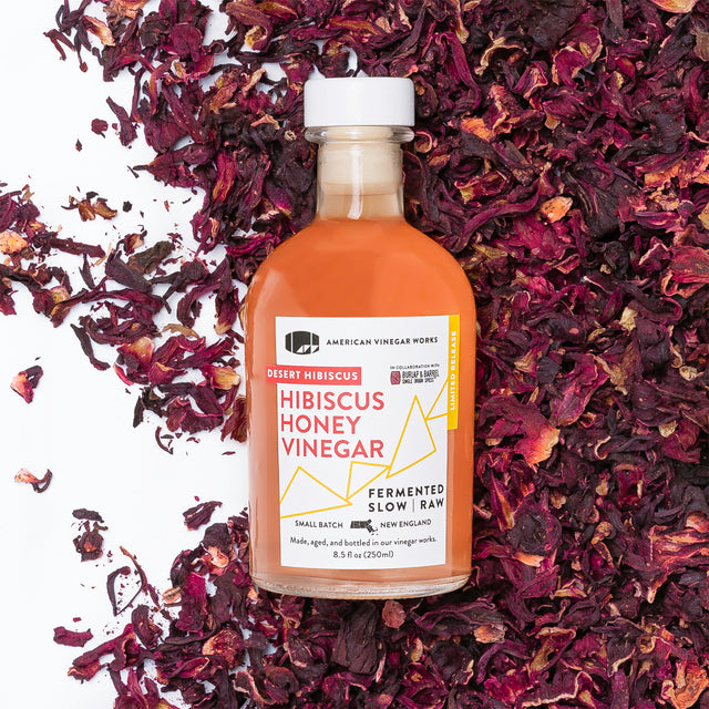 A bottle of Honey Hibiscus Vinegar with a label that reads 'Robert Hibiscus Honey Vinegar, American Craft Vinegar', surrounded by hibiscus flower petals.