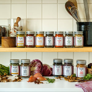 A set of 12 glass spice jars with silver lids and white labels from the brand 'Burlap & Barrel' arranged on two wooden kitchen shelves alongside various fresh and dried ingredients, set against a white tiled backsplash and light wood cabinets.