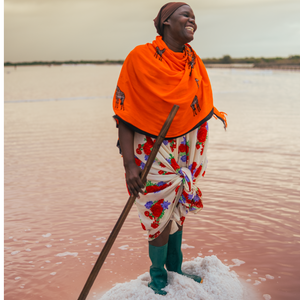 Partner farmer with an orange shawl smiling atop of a pile of salt. Surrounding her is water and she is holding a long stick.