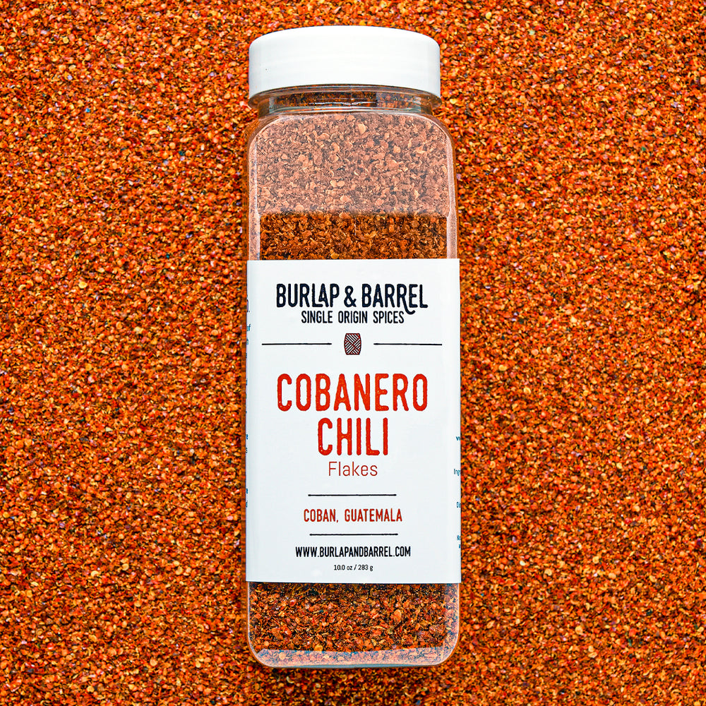 Container of cobanero chili flakes from burlap & barrel on a background of loose chili flakes.