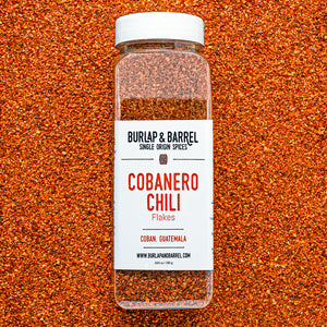 Container of cobanero chili flakes from burlap & barrel on a background of loose chili flakes.