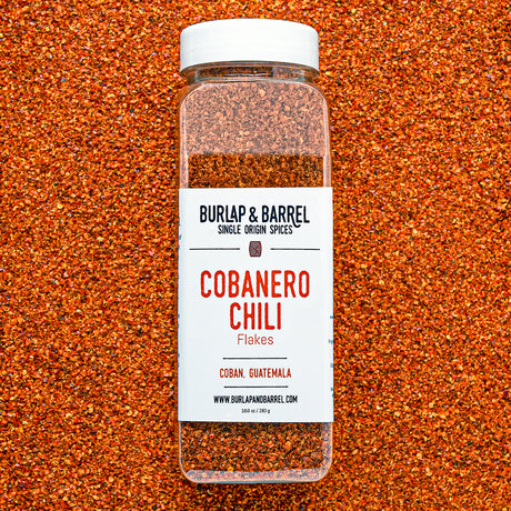 Container of cobanero chili flakes from burlap & barrel on a background of loose chili flakes.