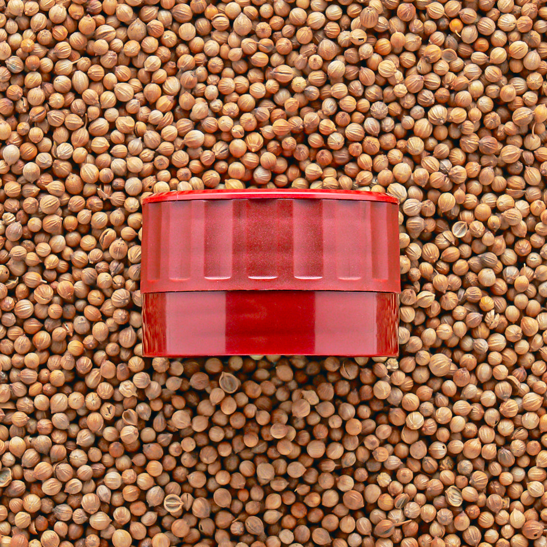A red adjustable grinder-top that goes onto Burlap & Barrel jars on a bed of seeds.