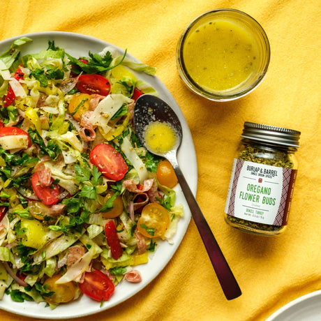 Salad with a jar of oregano flower buds and a small bowl of dressing on a yellow tablecloth.