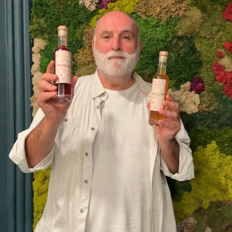 A man with a white beard and bald head holds up two bottles of syrup, one in each hand, in front of a green moss wall. He is wearing a white button-down shirt over a white t-shirt and black pants.