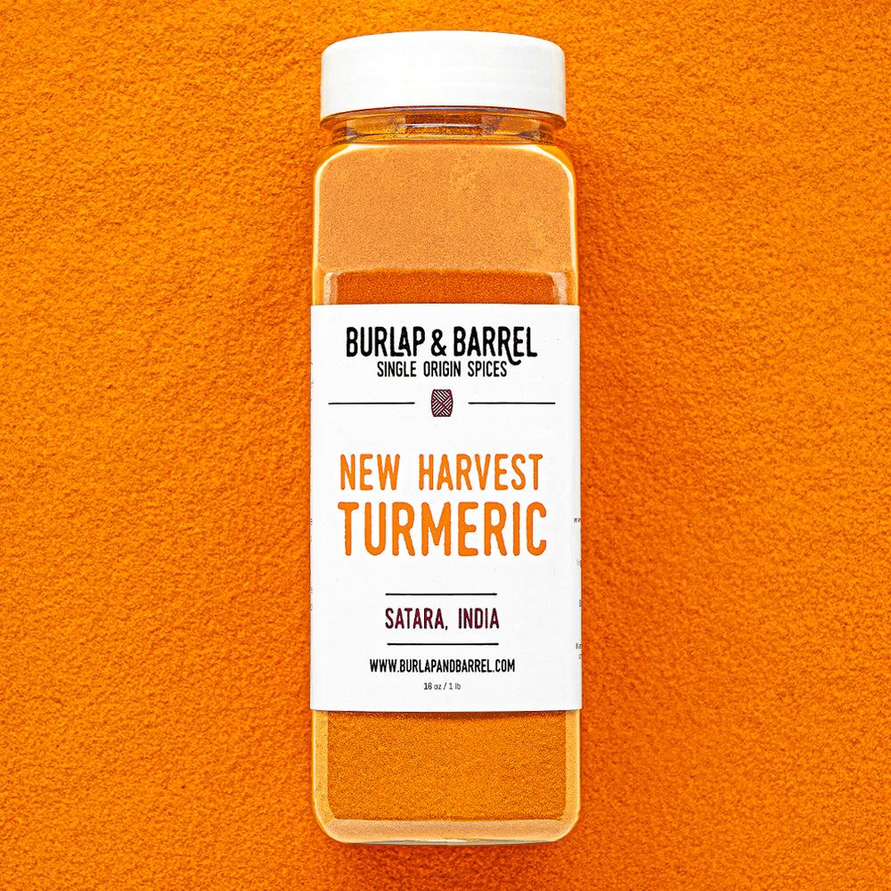 Plastic container of New Harvest Turmeric from Burlap & Barrel with a white lid on a background of loose turmeric powder.