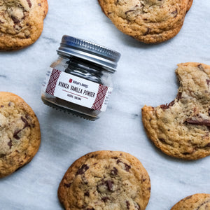 Jar of vanilla powder surrounded by chocolate chip cookies on a marble surface
