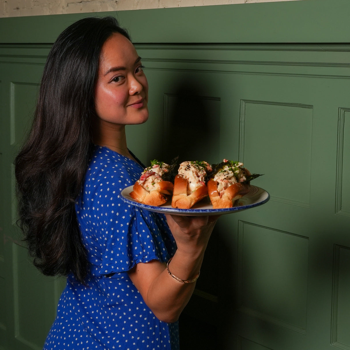Woman in a blue dress holding a plate of food against a green wall.