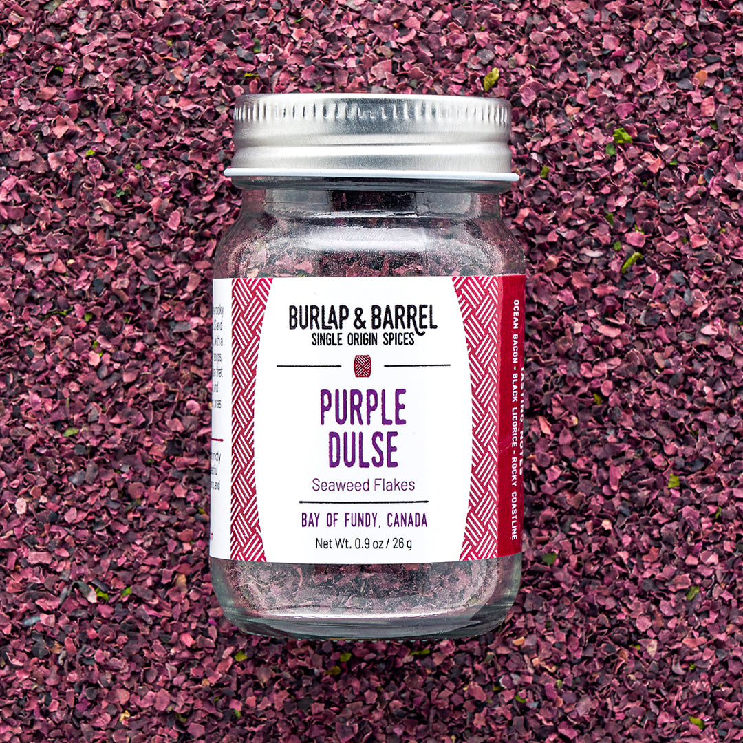 A jar of Purple Dulse Flakes by Burlap & Barrel, sustainably hand-harvested from the Bay of Fundy, Canada, displayed on a background of dulse flakes.