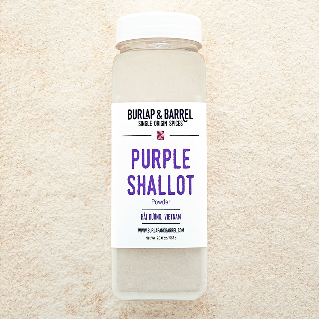 A container of Purple Shallot Powder by Burlap & Barrel, displayed on a background of beige powder.