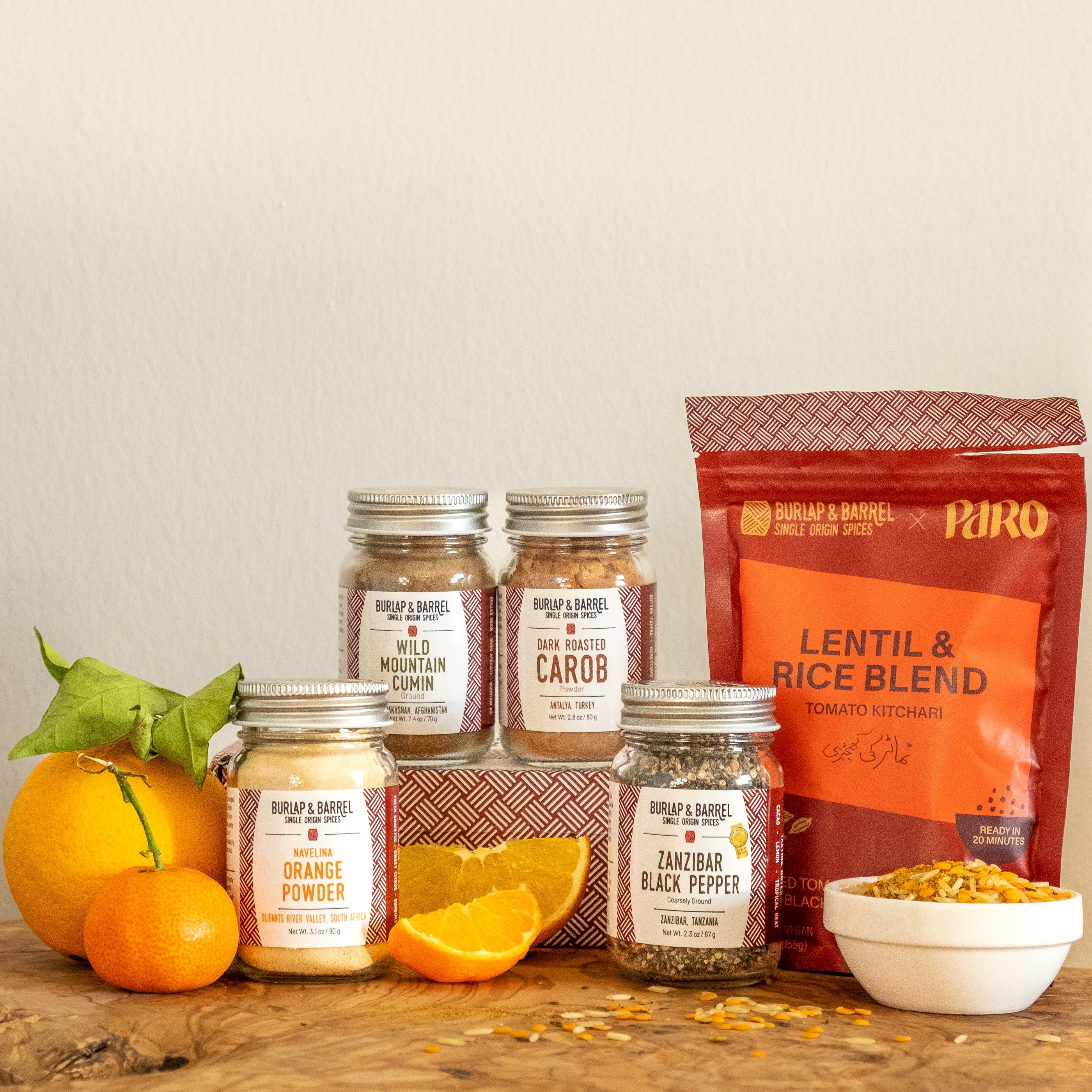 Jars of Spices displayed next to whole and sliced oranges with a bag of lentil & rice blend and a white bowl showing the lentil and rice. 