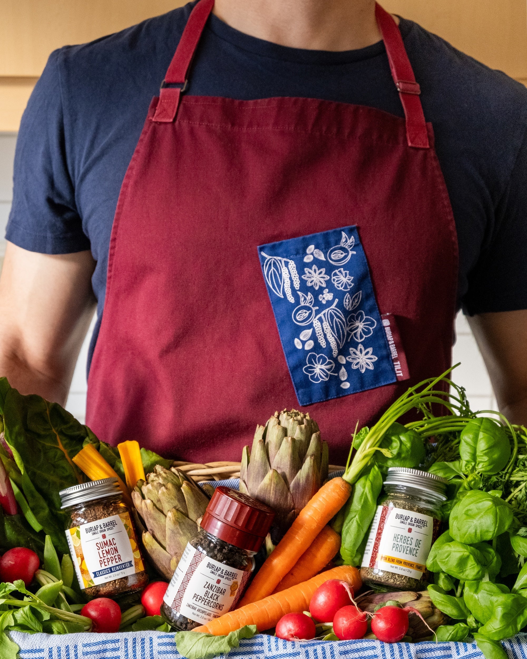 Person holding a basket of fresh vegetables and spices, wearing a red apron with a blue pocket.
