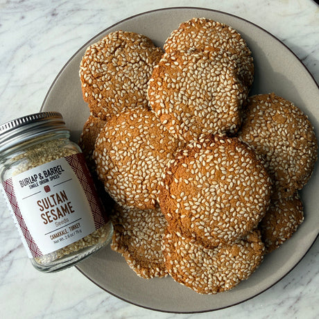 A plate of golden-brown sugar cookies heavily topped with white sesame seeds sits on a white marble surface next to a glass jar of Burlap & Barrel Seasoned Sesame.