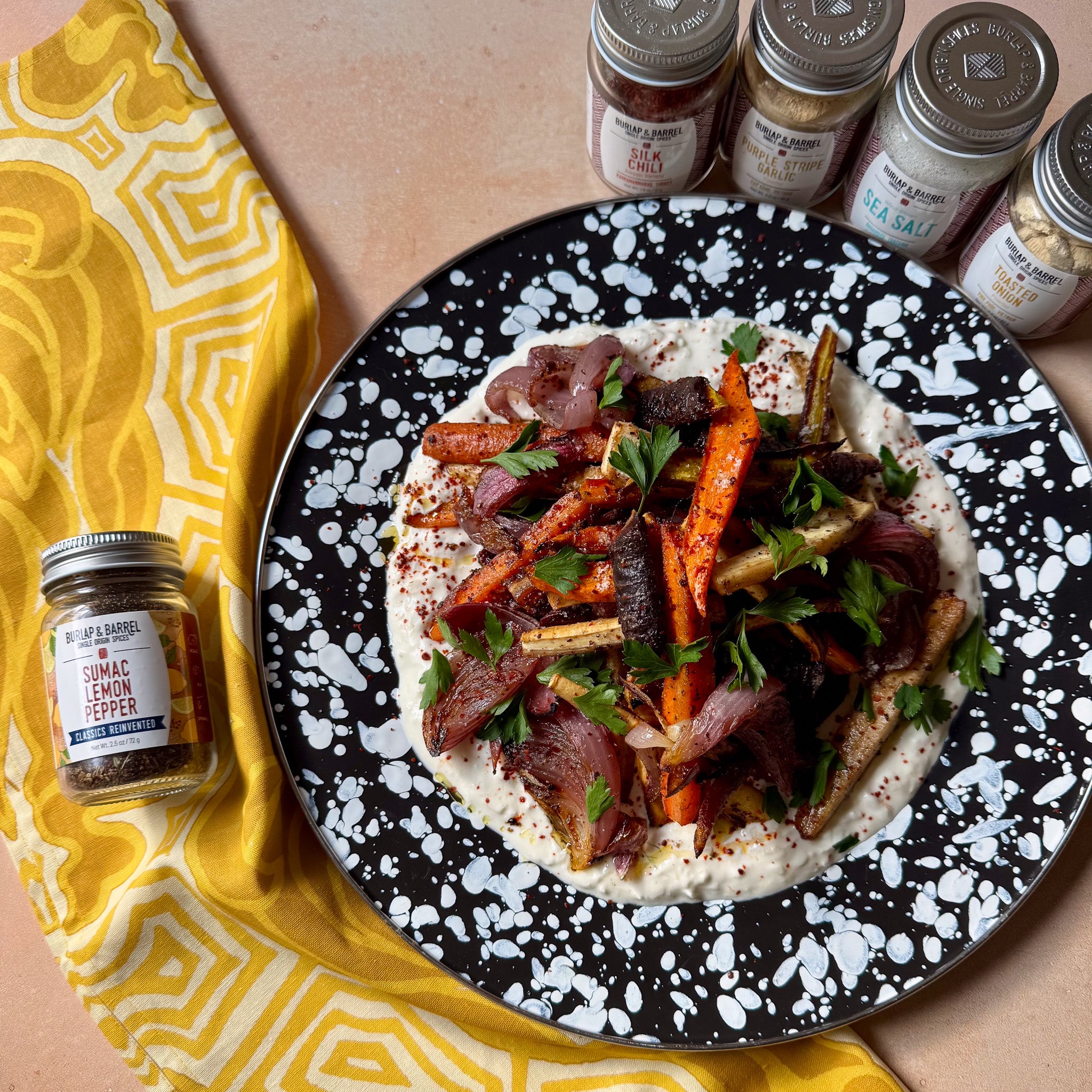 Plated dish with roasted vegetables and a black and white speckled plate, surrounded by Burlap & Barrel spice jars. 