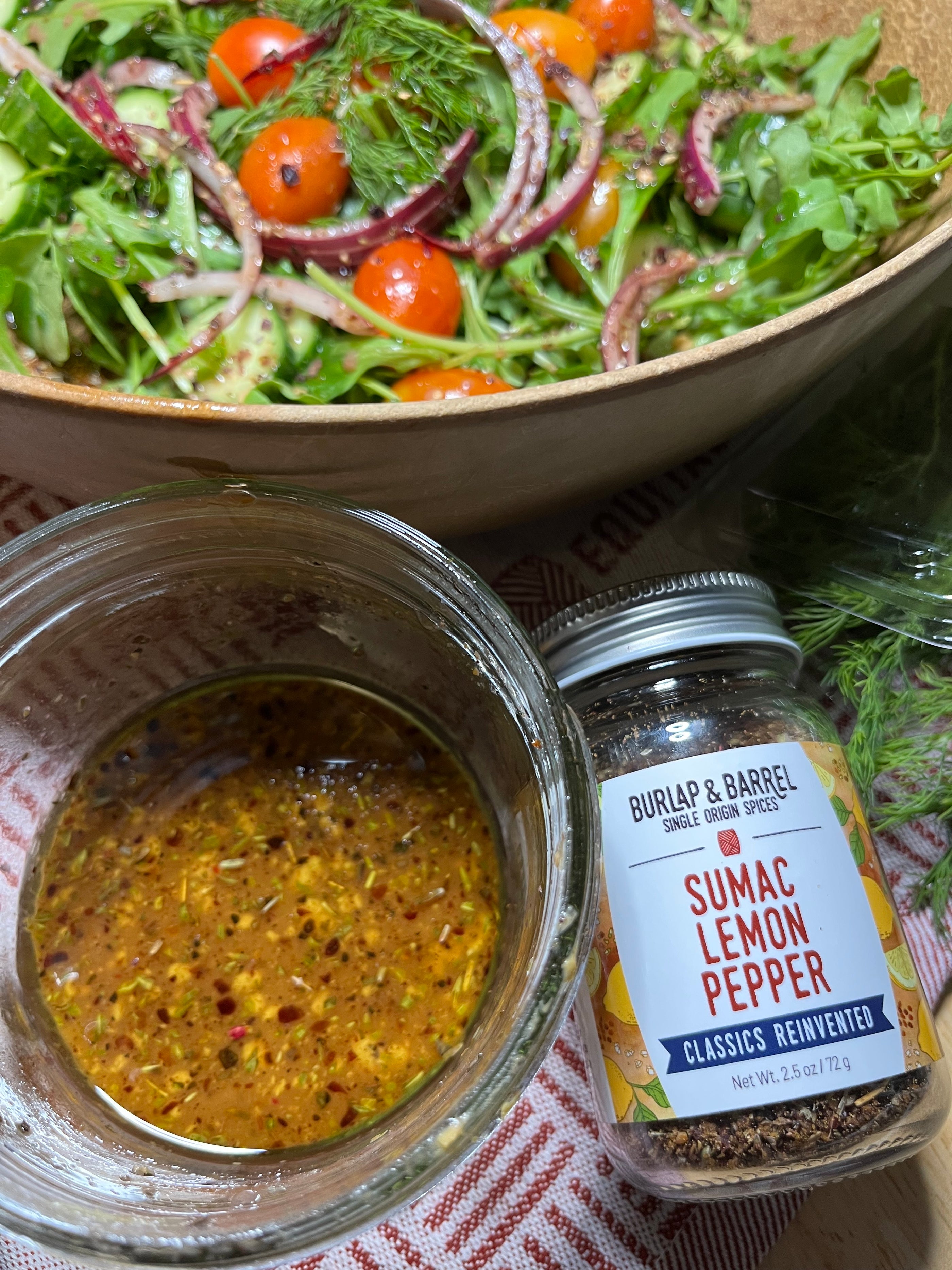 Jar of Sumac Lemon Pepper next to a green salad with sliced red onion and cherry tomatoes and a bowl of vinaigrette. 