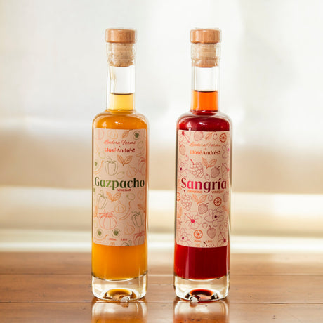 Two clear glass bottles of natural liquid nectaries with cork stoppers, standing on a wooden table in front of a softly blurred window. The left bottle is labeled Gazpacho with golden yellow liquid, and the right bottle is labeled Sangrie with deep red liquid.