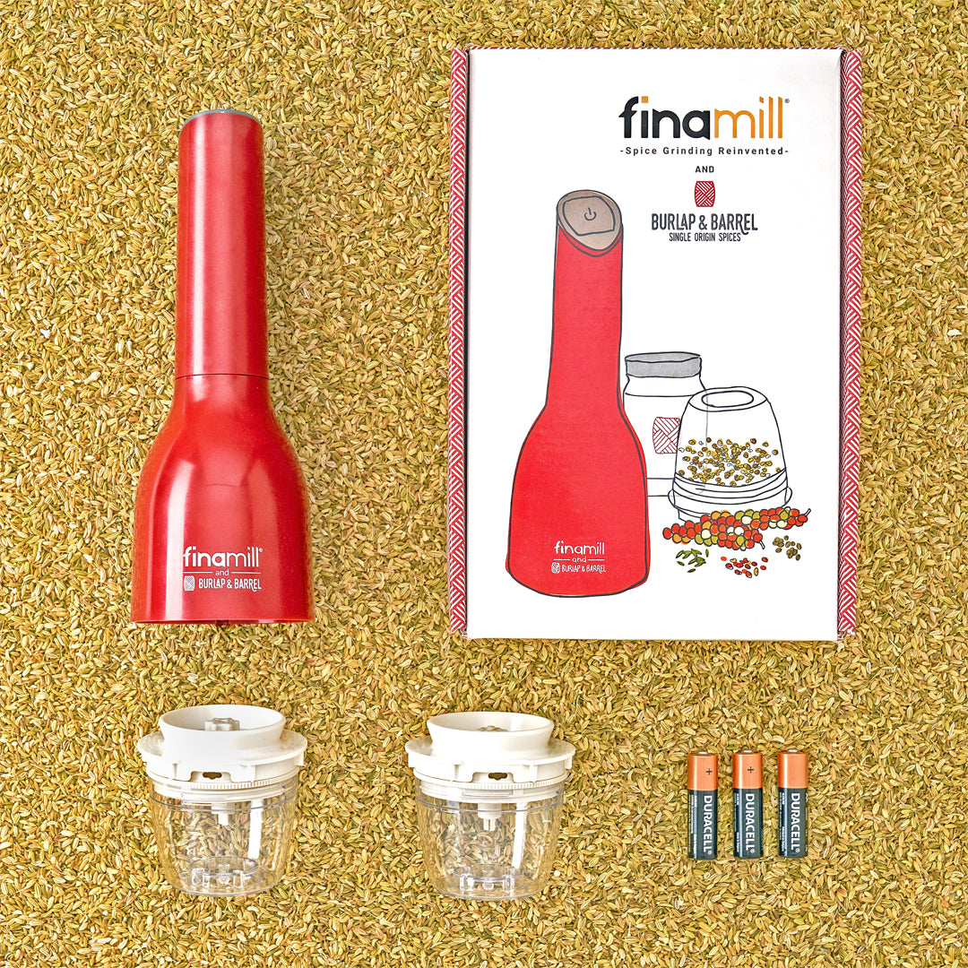 Finamill automatic grinder with the box it comes in, two removable grinding pods and 3 batteries on a bed of loose fennel seeds